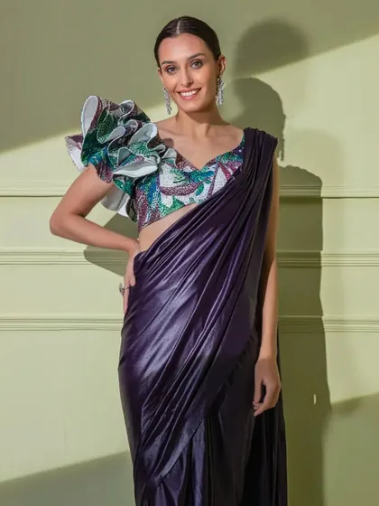 For the fashion-forward woman, this purple metallic drape saree is paired with a statement blouse featuring a dramatic, multi-layered ruffle on one shoulder.