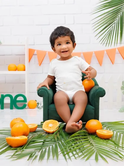 A happy baby girl sits in a miniature green armchair, surrounded by oranges and palm leaves for a fresh and fun theme.