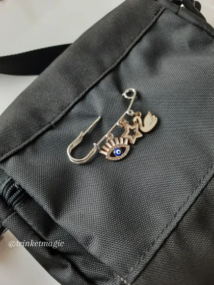 Pin your vibe to your bag or jeans with this safety pin charm. This one features a protective evil eye, a star, and a graceful swan in a silver and rose gold finish.