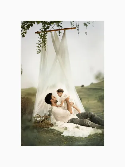 A father lies on a blanket in a field, lifting his baby daughter into the air. This playful and loving interaction, framed by a beautiful canopy, is a perfect capture of the father-daughter bond.
