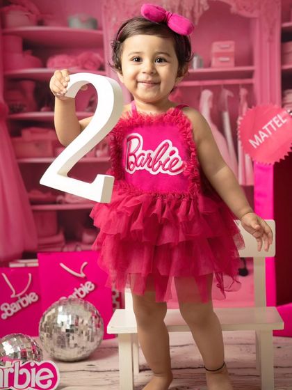 Celebrating turning two with a Barbie theme. This little girl proudly holds up the number '2' in her pink dress, surrounded by disco balls and Barbie accessories.