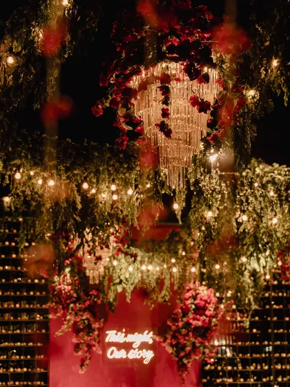 A stunning view of the Sangeet decor, with chandeliers dripping with red flowers and a neon sign glowing against a wall of candles. The layers of light and florals created a magical, immersive experience.