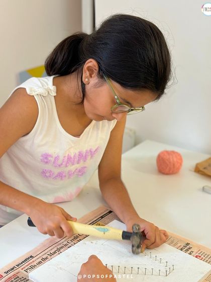 A young artist learns to use a hammer safely to place the nails for her initial string art project, developing new skills and concentration.
