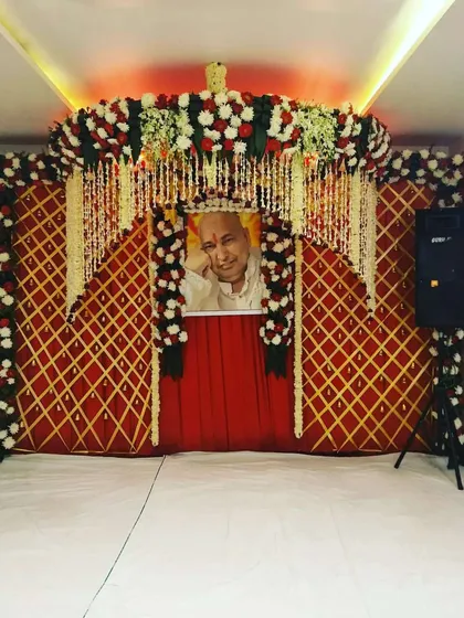 A classic darbar backdrop using a red and gold lattice screen. The portrait of Guru Ji is the centerpiece, framed by arches of red and white flowers with hanging floral strings.