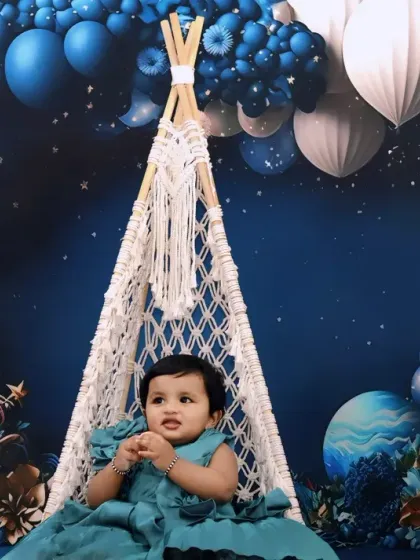 A baby girl sits in a macrame swing for her first birthday, surrounded by a stunning blue and white balloon arrangement and 'ONE' blocks.