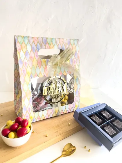 A pastel-colored Diwali gift bag with a window, revealing the treats inside. A box of four assorted chocolates is displayed alongside.