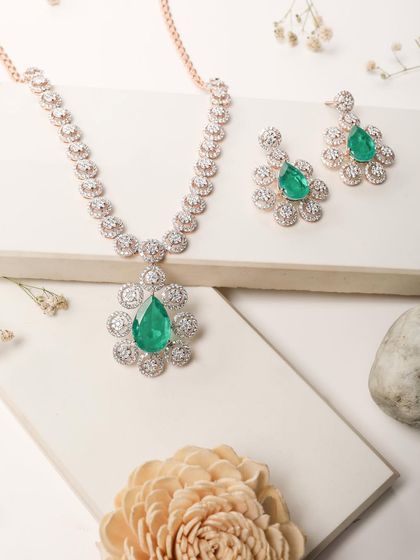 A stunning emerald and diamond necklace set, beautifully styled on a white block with delicate flowers. This clean and elegant composition highlights the luxury of the piece.