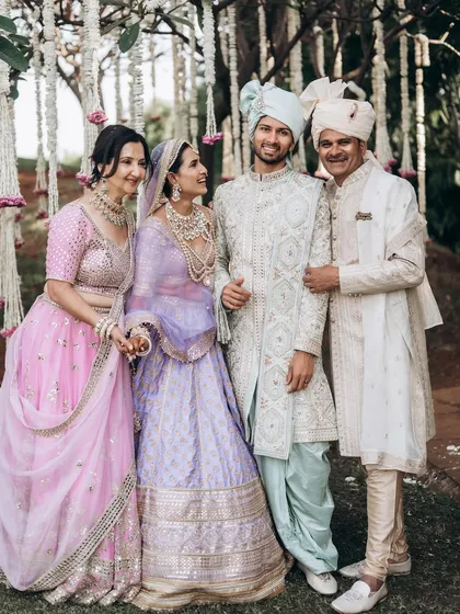 A bride and groom pose with their parents under a beautiful floral arrangement. The coordinated pastel outfits and genuine smiles make this a perfect multi-generational family portrait from a wedding day.