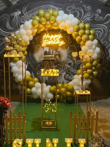 An outdoor party setup with a white and gold balloon ring, neon sign, and elegant gold-toned props.
