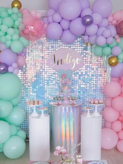 This mermaid theme features a gorgeous pastel balloon garland over an iridescent shimmer wall. The setup includes holographic and clear pedestals for the cake and desserts, adding to the magical, under-the-sea vibe.