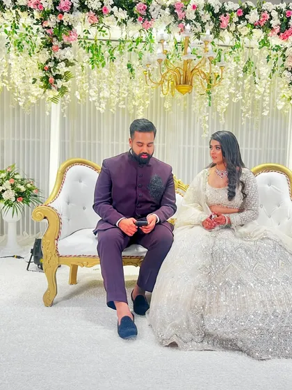 The happy couple seated on a classic white and gold stage after their grand entrance. The backdrop of hanging white florals adds a touch of romance and elegance.