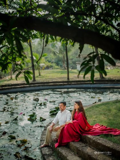 A serene scene by a lily pond. The couple sits together, the bride's red saree flowing beautifully, creating a peaceful and romantic image.