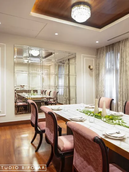 The formal dining room at the Wanderlust Villa is bright and reflective, with a large mirrored wall that enhances the sense of space. Dusty rose chairs add a soft touch of color against the white marble table.