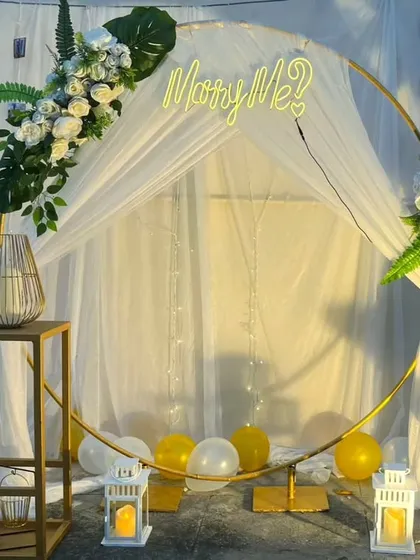 A modern, bohemian-style proposal backdrop. A large golden ring is draped with white fabric and accented with white florals and pampas grass, complemented by a "Marry Me?" neon sign for a trendy and romantic look.