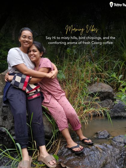 Morning vibes in Coorg. Two women share a warm hug by a stream, ready to start their day with misty hills and the aroma of fresh coffee.