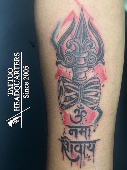 A modern take on a spiritual tattoo, this design features a Trishul and Damaru with "Om Namah Shivaya" in Devanagari script, accented with a splash of color.