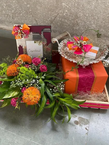 From welcome gifts to wedding favors, our curated Panos hampers add a sparkle to any celebration. This one features a silver tray and a gift box, surrounded by a lush arrangement of orange and green florals.