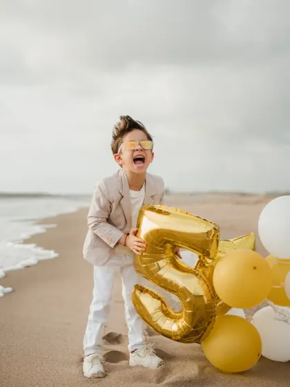 The birthday boy laughs with joy, holding a giant number "5" balloon on the beach. This shot perfectly captures the excitement of turning five.