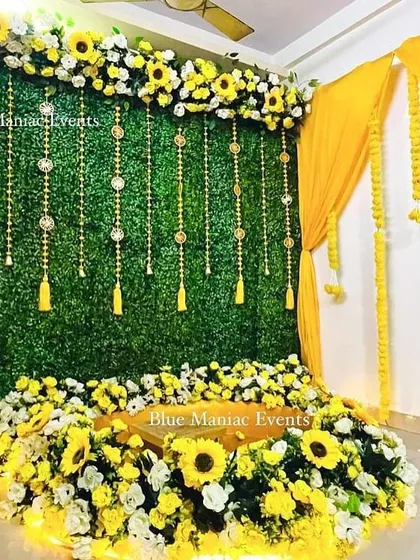A Haldi and Mehendi setup featuring a green wall with hanging tassels and a base of sunflowers. The yellow drapes and marigold garlands add a traditional touch.