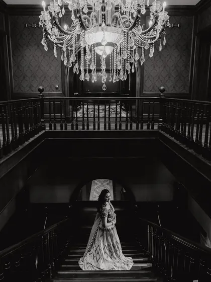 Another artistic black and white shot from the Sangeet. This full-length portrait captures the bride's elegance against the dark, moody backdrop of the grand staircase, creating a feeling of timeless romance.