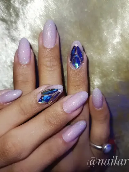 A beautiful set of almond-shaped nails with a soft lavender glitter base and large, eye-catching blue crystal accents.