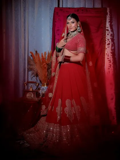 The bride poses confidently in her red lehenga, showcasing the full ensemble against a simple red backdrop.