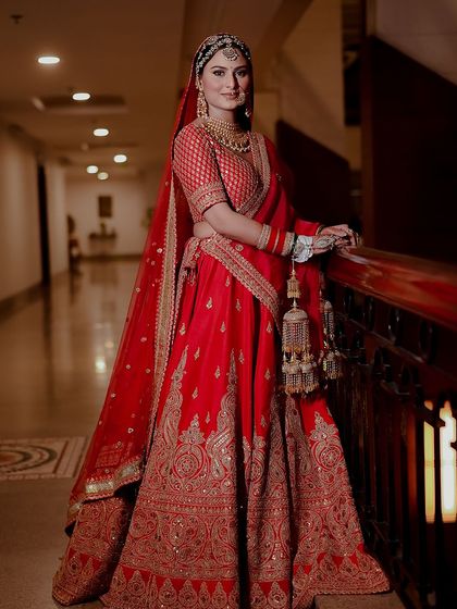 A full-length bridal portrait showcasing a stunning red lehenga with intricate gold embroidery. The bride's confident and graceful pose makes this a powerful and elegant image.