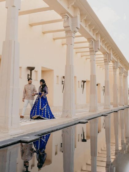 A stunning reflection shot at The Oberoi Udaivilas. The couple walks along the edge of a water body, their royal blue and cream outfits perfectly mirrored in the still water, creating a symmetrical and visually arresting image.