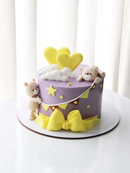 A lovely baby shower cake in shades of purple and yellow, decorated with cute teddies and a big bow.