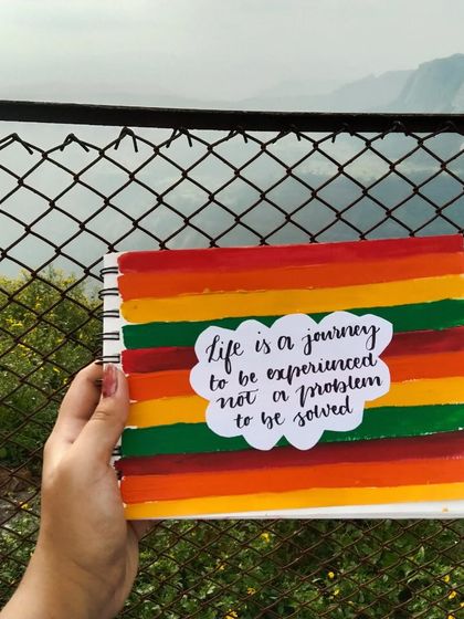 A colorful, striped painting in my sketchbook with the quote "life is a journey to be experienced, not a problem to be solved," held up against a scenic mountain view.