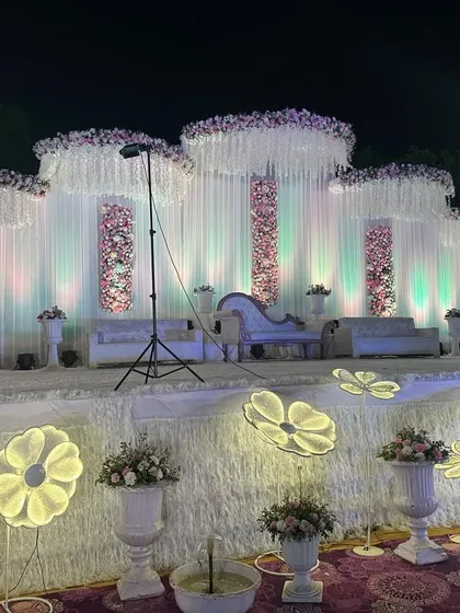 An elegant, all-white wedding stage design that feels fresh and sophisticated. The backdrop uses flowing white drapes and panels of pink and white flowers, with unique illuminated floral lights in the foreground.