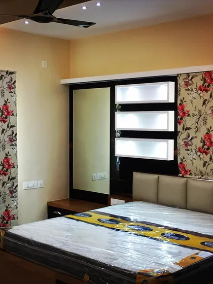 A bedroom in a Bangalore flat featuring a classic floral wallpaper that doubles as a window curtain. The design incorporates a simple, clean-lined bed and a wardrobe with an integrated dressing mirror.