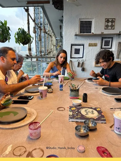A family sits around the large pottery table, focused on painting their plates, with the beautiful city view visible from the open balcony.