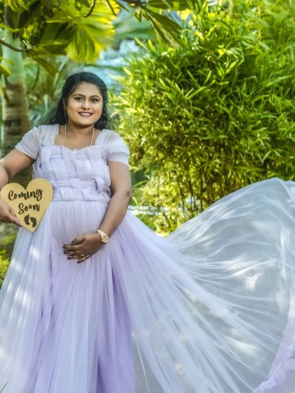 A beautiful outdoor solo portrait holding a "Coming Soon" sign. The flowing lavender gown and the lush green background create a soft and dreamy pregnancy announcement.