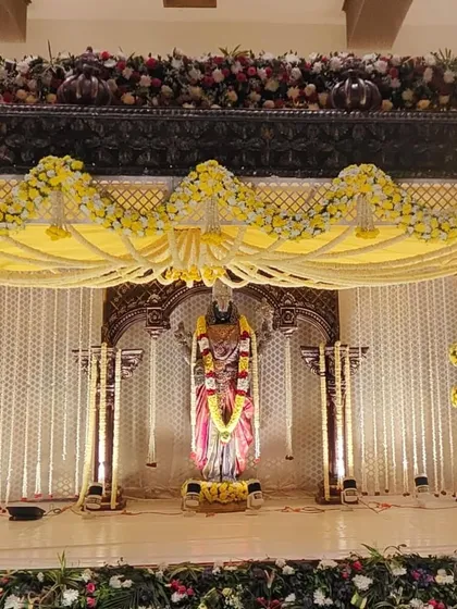 This is a traditional South Indian wedding mandap I built around a central deity statue. The design uses yellow and white fresh flowers for the garlands and pillar decorations, set against a classic carved wooden frame.