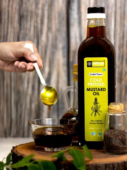 A hand holds a spoonful of golden mustard oil, with the drip captured mid-air. This action shot emphasizes the oil's rich color and viscosity, set against a rustic, traditional backdrop.