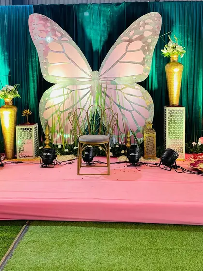 A whimsical stage setup for a baby shower, featuring a giant pink and iridescent butterfly as the main backdrop. The stage is decorated with golden vases and floral arrangements.