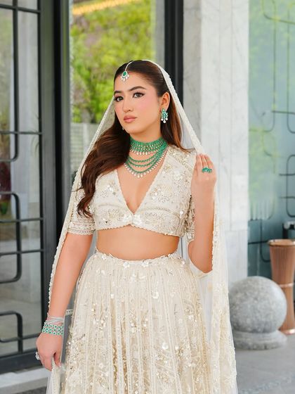The full bridal look, showcasing the modern ivory lehenga paired with a statement emerald choker. The makeup is kept soft and radiant to maintain a fresh, contemporary feel.