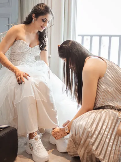 A bridesmaid helps the bride with her stylish white sneakers, a perfect blend of elegance and comfort. It's these small, supportive moments with the bride tribe that make the getting-ready process so special.