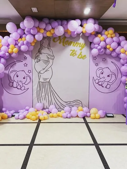 A sweet baby shower setup in a lavender and yellow color scheme. The backdrop features a 'Mommy to be' silhouette and cute elephant illustrations, framed by a balloon arch.