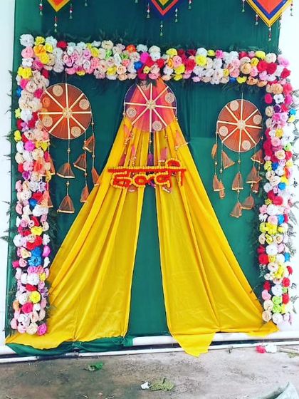 A beautiful Haldi backdrop featuring a floral arch and traditional dreamcatcher hangings against a yellow and green curtain.