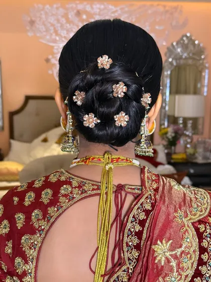 The back of a sleek bridal bun, dotted with delicate gold flower pins that match the embroidery on the bride's red lehenga.