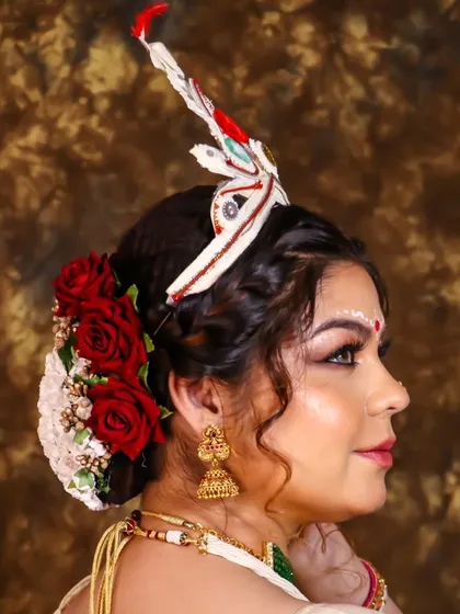 The side profile of a traditional Bengali bridal hairstyle, featuring a classic bun adorned with red roses and the shola mukut.