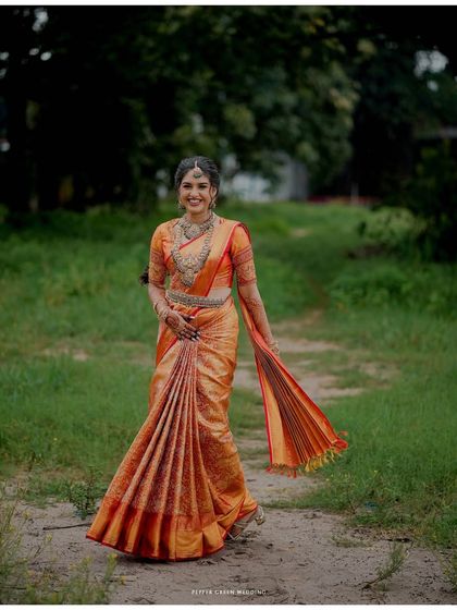 A full-length portrait of the bride in her stunning orange Kanjivaram saree, her joyful expression lighting up the frame as she walks through a green field.