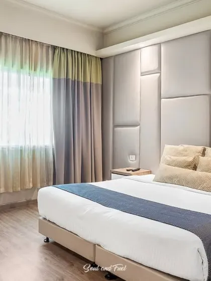 A wide shot of a modern hotel room at Sinclairs, Ooty, with a large, comfortable bed and a padded feature wall. The neutral color palette creates a calm and restful environment for travelers.