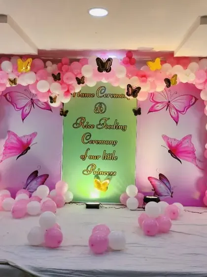 A sweet and simple butterfly theme for a naming and rice feeding ceremony. The backdrop is a soft green and pink, framed by a pink and white balloon arch with gold and black butterfly accents. This is a great example of a beautiful setup for a smaller stage or venue.