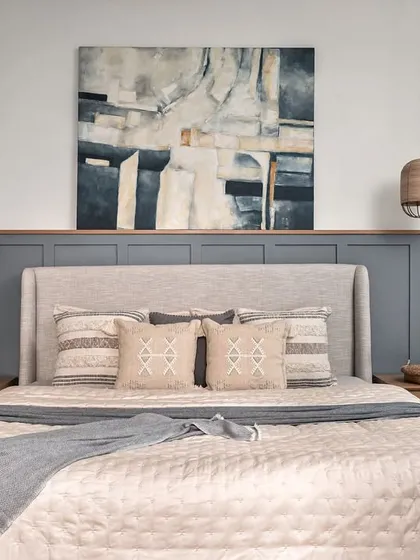 A master bedroom should be a true retreat. Here, the design is centered around tranquility, with a light beige upholstered bed set against soothing grey wainscoting. The abstract canvas and oak side tables complete this modern farmhouse bedroom, creating a perfect balance of comfort and rustic elegance.