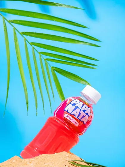 A tropical-themed shot of a Papa Nata strawberry drink, styled with sand and a palm leaf. This creates a vacation vibe, associating the beverage with refreshment and fun.