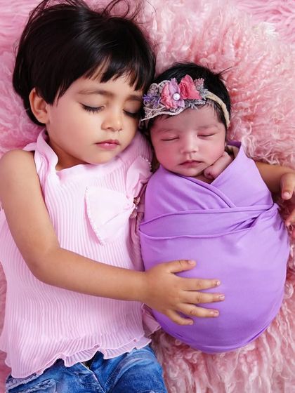 A heart-melting moment of pure love as an older sister sleeps beside her swaddled newborn sibling on a soft pink blanket.