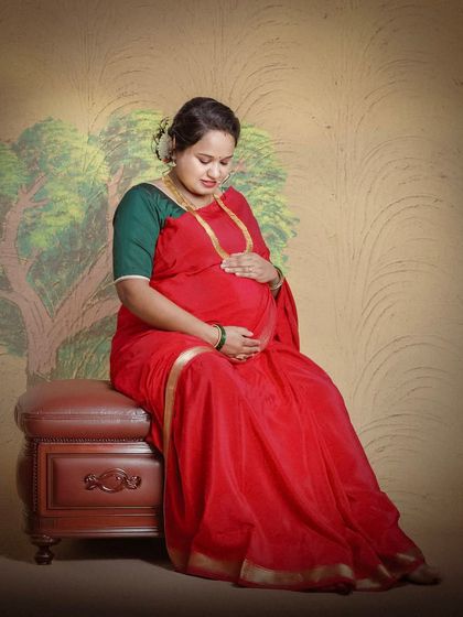 A serene portrait of a mother-to-be in a classic red saree. This studio shot uses a painted backdrop and soft lighting to create a warm, traditional feel.